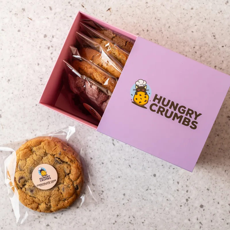 An open Hungry Crumbs box of individually wrapped cookies, with one wrapped chocolate chip cookie beside it