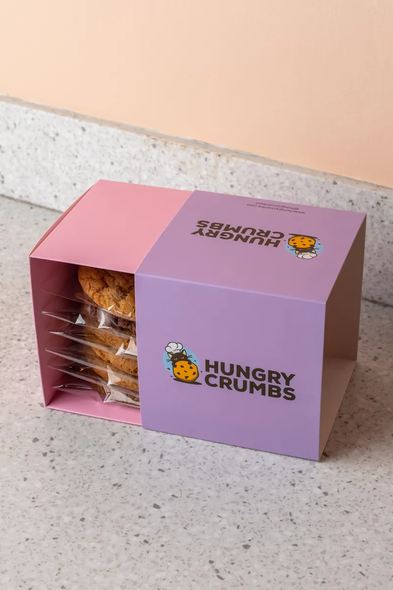 A lilac Hungry Crumbs box, open at one end to show wrapped cookies, against a terracotta wall