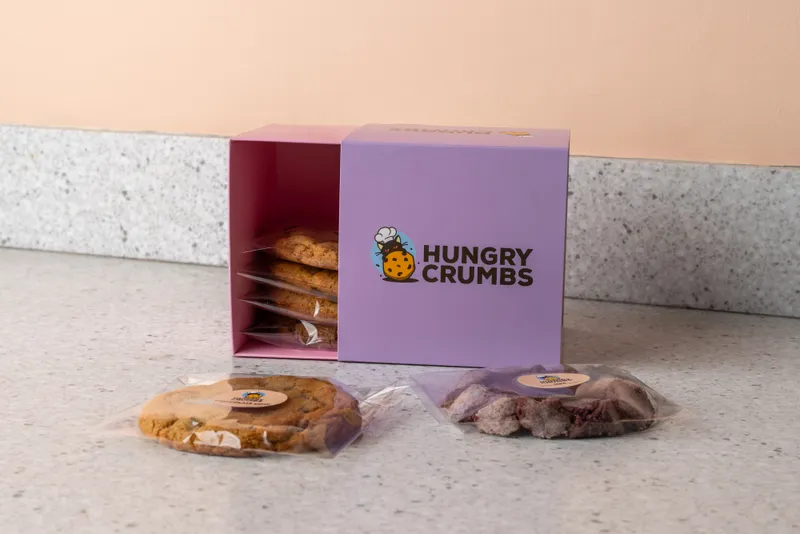A lilac Hungry Crumbs box with the cat-and-cookie logo, open, with individually wrapped cookies inside