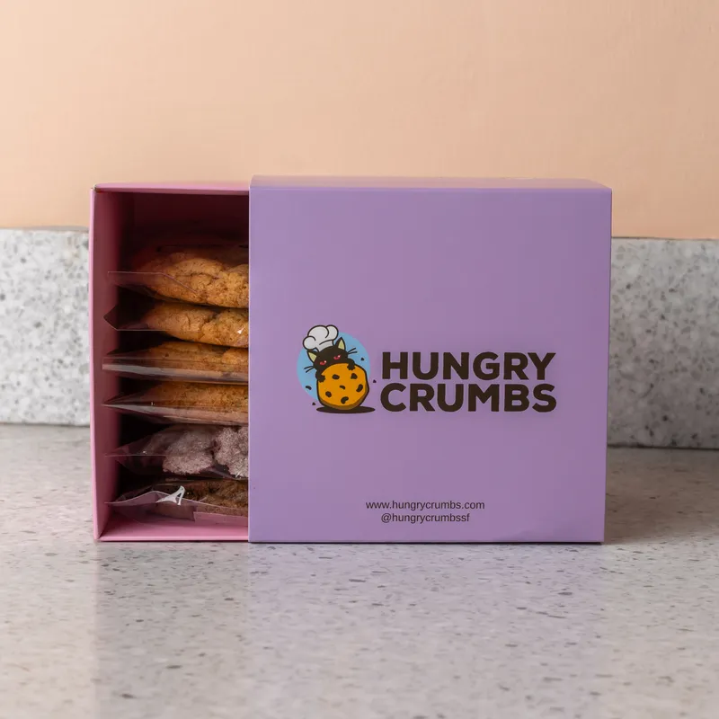 A lilac Hungry Crumbs box with its sleeve slid back, individually wrapped cookies standing on edge inside, against a terracotta wall