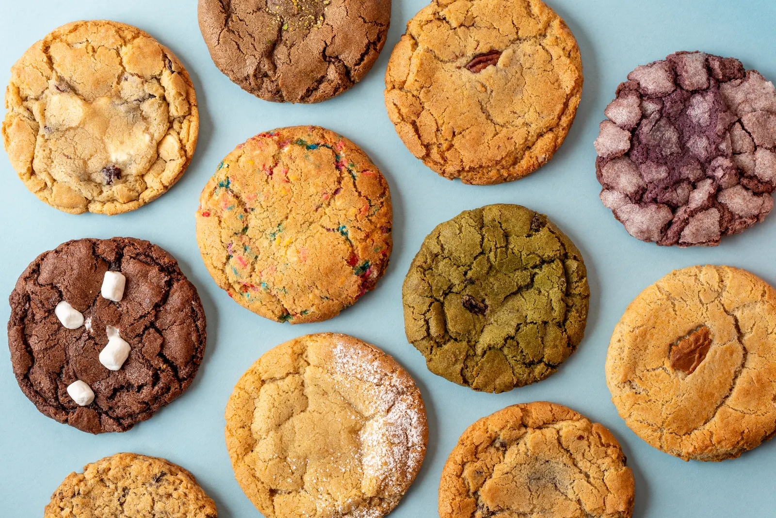 Hungry Crumbs cookies edge to edge on a pale blue backdrop — funfetti, matcha, an ube crinkle, pecan and a chocolate one topped with marshmallows among them