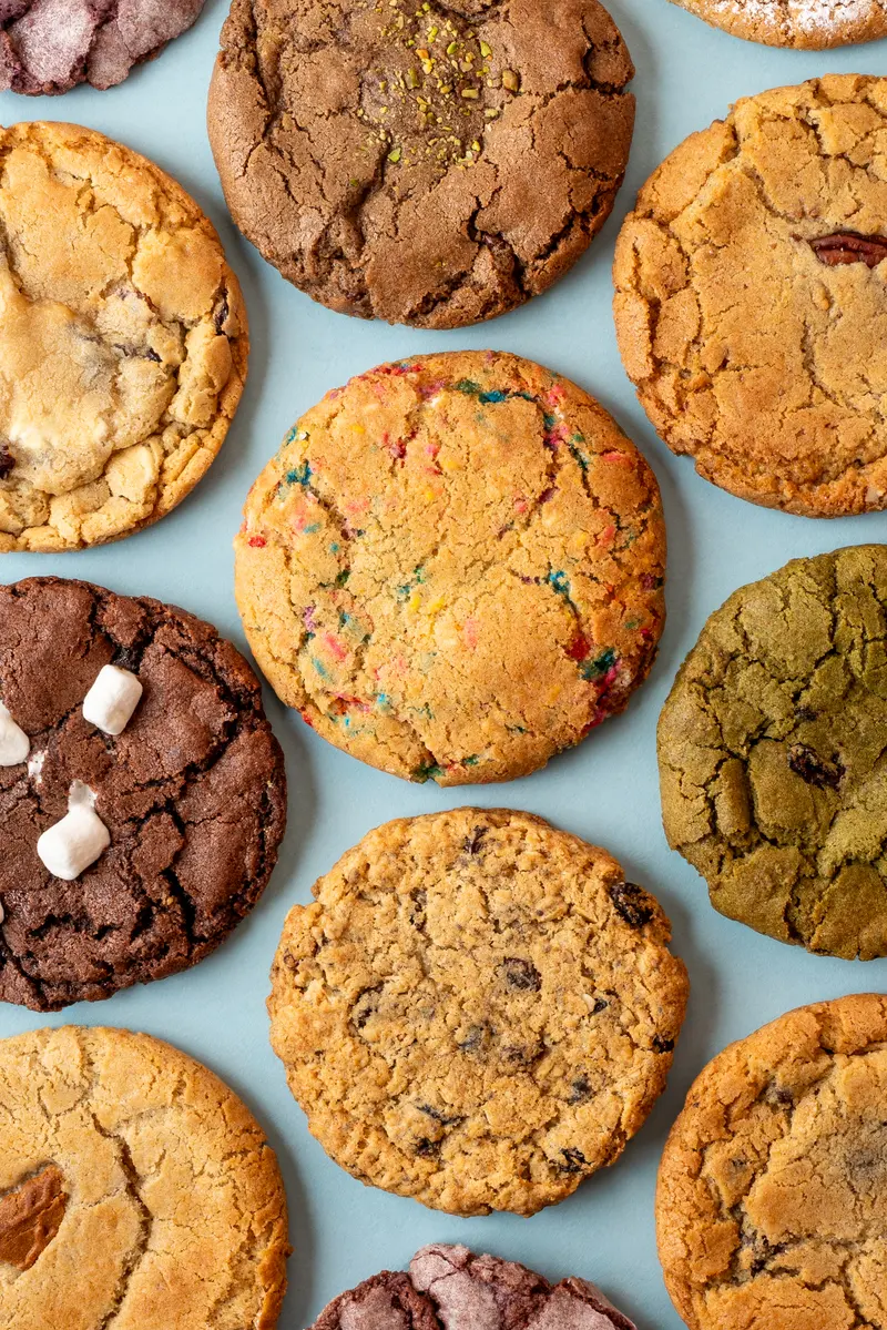 Hungry Crumbs cookies laid out on a pale blue backdrop — sprinkle, matcha, oatmeal raisin and a chocolate one topped with marshmallows among them
