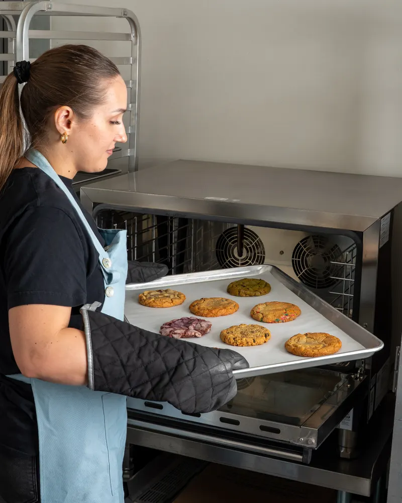 A baker in a blue apron and oven gloves sliding a tray of seven just-baked cookies out of the oven in the Hungry Crumbs kitchen