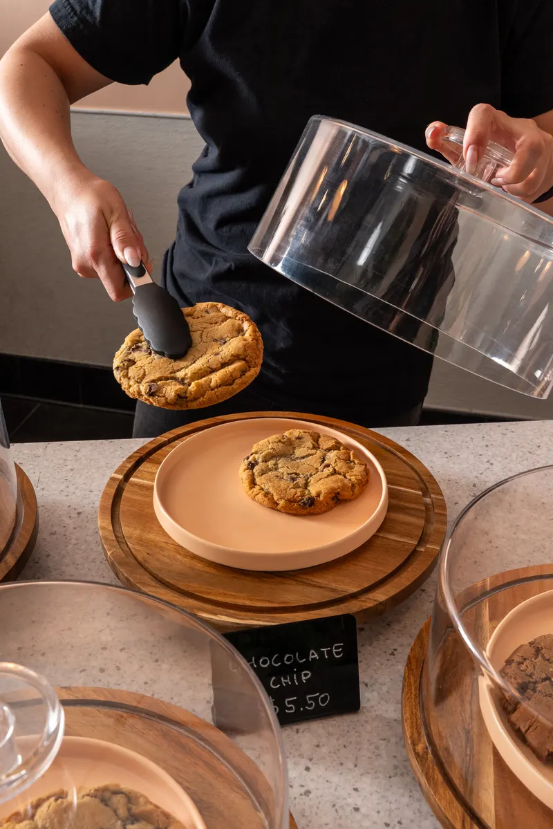 A cookie lifted with tongs from under its dome onto a pale pink plate at the counter