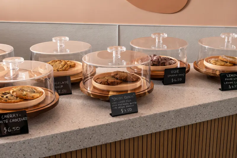 A row of glass cake domes along the counter edge, each holding cookies on a wooden board with a hand-chalked name tag