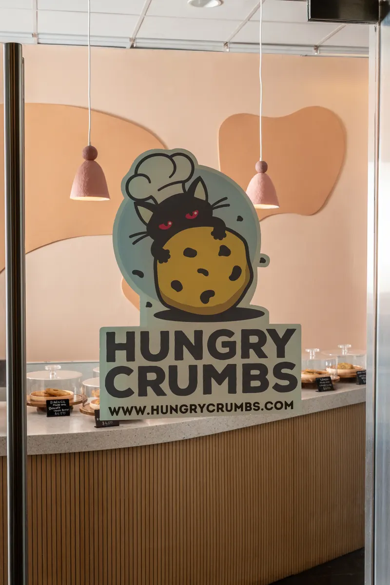 The Hungry Crumbs decal on the glass door — a cat in a chef's hat leaning over a chocolate chip cookie, www.hungrycrumbs.com beneath — with pendant lamps and the counter behind