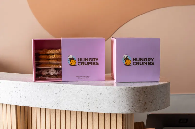 Two lilac Hungry Crumbs boxes on the shop's curved terrazzo counter, the left one open to show wrapped cookies stacked inside, above the fluted oak counter front and below a terracotta shape on the peach wall