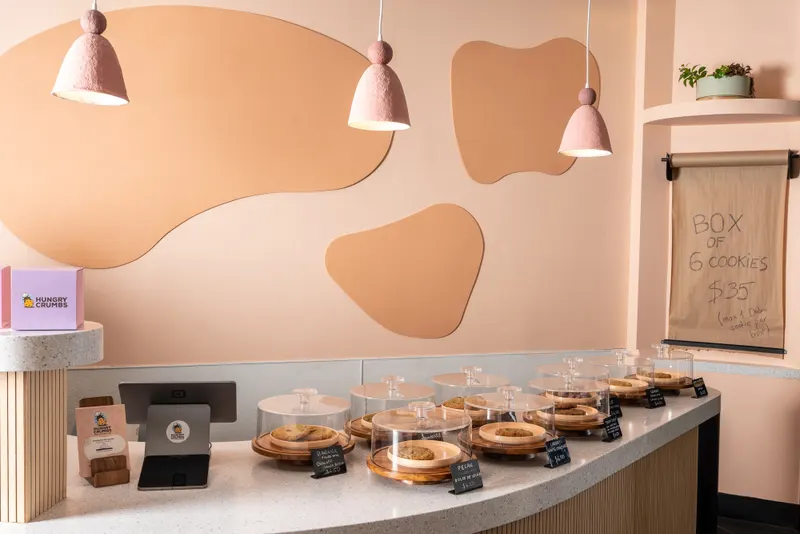 The counter from the register end — cookie domes down the terrazzo curve, terracotta pendants above, and a kraft-paper sign on the wall reading "Box of 6 cookies $35"