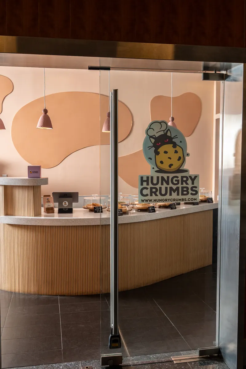 The glass entrance door seen straight on, the Hungry Crumbs decal at eye height and the curved wooden counter with cookie domes inside